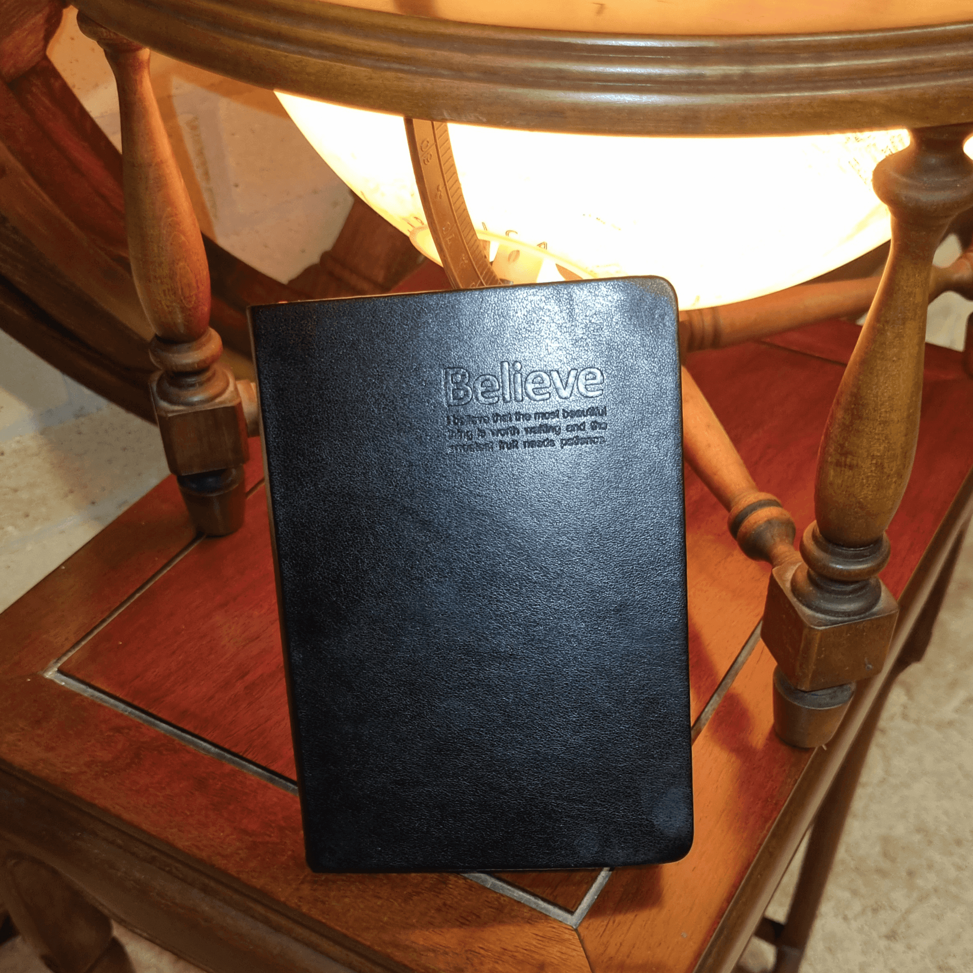Black Genuine Leather Gold Edged Travel Journal with 'Believe' embossing on a wooden table near a globe.