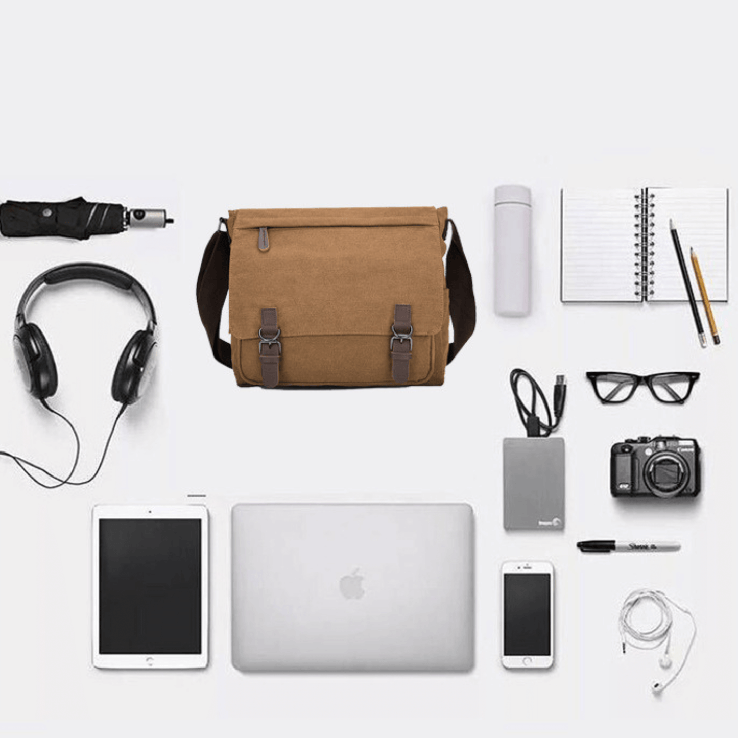 Brown canvas cross body messenger satchel bag surrounded by tech gadgets and office supplies.